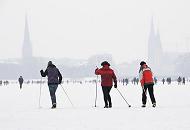 245_6109 Skilangluferinnen nutzen den hohen Schnee, der auf dem Alstereis der Aussenalster liegt, um eine Runde um den 164 ha groen Hamburger Binnensee zu machen; die Rundstrecke am Alsterufer ist fr Jogger ca. 7,5 km lang. Im Hintergrund in den Schneewolken die Kirchtrme der St. Petri und Nikolaikirche sowie rechts der Rathausturm.  