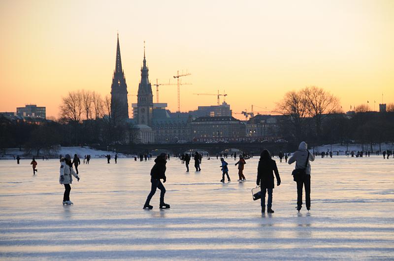 Alstereisvergngen auf der Hamburger Auenalster - Sonnenuntergang. 254_5925 Bis in den Abend hinein nutzen die Hamburger und Hamburgerinnen die Mglichkeit auf der zugefrorenen Aussenalster Schlittschuh zu laufen oder einfach nur spazieren zu gehen. Ein Zug fhrt gerade ber die Lombardsbrcken - im Sonnenuntergang zeichnen sich die Baukrne und der Turm des Rathauses und der Nikolaikirche im roten Abendhimmel ab.