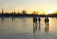 257_5875 Spaziergänger dem Eis der zugefrorenen Alster gehen Richtung Hamburg - St. Georg. Das Licht der untergehenden Sonne spiegelt sich auf der blanken Eisdecke.