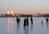 259_5885 Die untergehende Sonne stahlt die Hochhäuser an der Mundsburg an - das Licht spiegelt sich auf dem Eis der zugefrorenen Alster. Zwei Schlittschuhläufer spielen Eishockey, andere gehen gegen die Kälte dick mit Schal und Mütze eingemummt auf dem Alstereis spazieren.