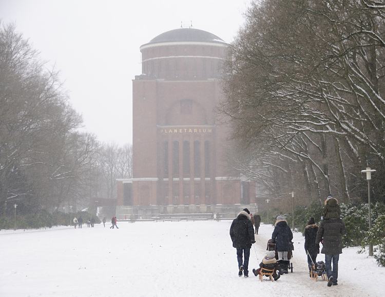 Familienspaziergang mit Schlitten im Stadtpark Hamburg Winterhude. 264_5083 Familien gehen im Schnee des Hamburger Stadtparks spazieren - Vter ziehen ihre Sprsslinge auf dem Schlitten oder tragen sie Huckepack, eine Mutter schiebt ihren Kinderwagen durch den Schnee auf dem Weg zum Planetarium. Der Schriftzug steht in goldfarbenen Buchstaben an der Fasse des 78m hohen Gebudes.