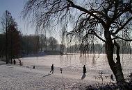 280_1010042 Der Jogger, der an einem Wintermorgen um den See des Hamburger Stadtparks joggt, wirft einen langen Schatten auf den Schnee. Ein anderer Stadtparkbesucher steht in der Sonne am Ufer und genießt den Blick auf den zugefrorenen See in der Sonne.