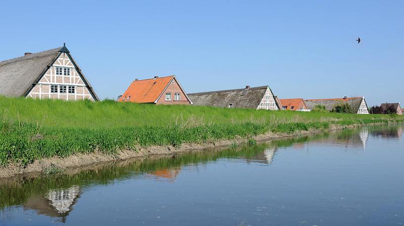 Bilder vom Alten Land; Hamburger Ausflugsziel am Wochenende. Fachwerkhäuser hinter dem Deich an der Lühe. 021_4078 Bauernhäuser und Wohnhäuser sind mit einem Deich vor der Überflutung durch Hochwasser der Lühe geschützt. Historische reetgedeckte Fachwerkhäuser und Klinkerhäuser stehen entlang des 12,7 km langen Flusses des Alten Landes. Die Giebel der Häuser spiegeln sich im ruhigen Wasser.