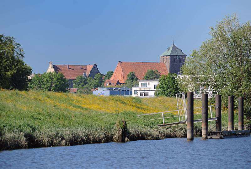 Schwinge, Stadt Stade - St. Wilhadi Kirche. Aufnahmer der Sehenswürdigkeiten der Metropolregion Hamburg. 073_4791 Blick vom Flusslauf der Schwinge zur Stadt Stade; alte Eisenpoller am Ufer der Schwinge zeugen von dem ehemaligen Wirtschaftsverkehr, der auf der Fluss stattgefunden hat. Am Deich des Flusses blühen gelbe Blumen - im Hintergrund die dreischiffige gotische Hallenkirche St. Wilhadi.
