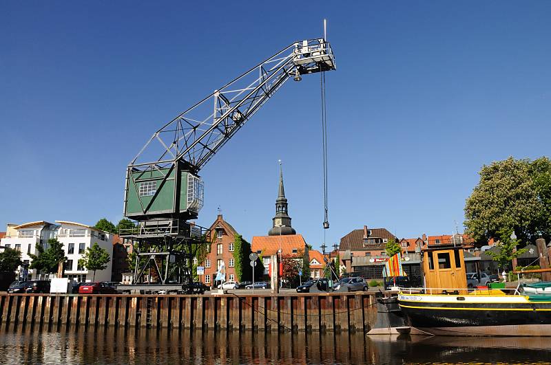 Kran am Ufer des Stader Hafens, Traditionsschiff Dora von Stade - Wohnhäuser + Kirchturm. Tourismus in der Metropolregion Hamburg / Hansestadt Stade. 083_8073 Hafenkai von Stadt, ein Rollkran steht am Hafenrand; im Hintergrund Wohnhäuser und ehemalige Speichergebäude. Rechts das Heck mit der Ruderanlage des 1902 in Itzehoe gebauten und jetzt restaurierten Küstenfrachters "DORA VON STADE".