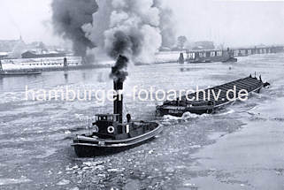 079_33_18 Ein Schlepper fhrt unter Dampf im Treibeis auf der Elbe - das Arbeitsschiff mit hohem Schornstein zieht  eine abgedeckte Schute, zwei Schiffer stehen am Heck des Kahns. Am Ufer sind die Zollanlagen von Hamburg Entenwerder zu erkennen.