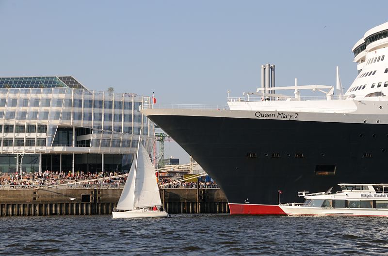 Schiffsbilder aus dem Hafen. Bug der Queen Mary 2 beim Cruise Center Hamburg / Chicago Kai, Schaulustige. Hamburger Hafenfotografie -  24_4828 Die Queen Mary 2 hat am Chicago-Kai des Hamburger Kreuzfahrtterminals festgemacht - Schaulustige sitzen auf der Terrasse und bewundern das grosse Schiff. Ein Segelboot kreuzt vor dem Bug des Luxusliners, ein Fahrgastschiff mit Touristen an Bord fahren an der Lngsseite des Schiffs entlang.  www.fotograf- hamburg.de