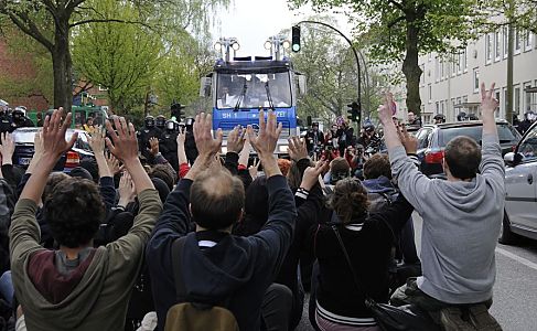 011_17487 - Antifaschisten und Anwohner des Hamburger Stadteils Barmbek versuchen mit gewaltfreien Strassenblockaden den Marsch der Neonazis zu verhindern. Hier heben die Blockierer zum Zeichen der Gewaltfreiheit ihre Hnde - der Wasserwerfer fhrt auf, von Seiten der Polizei wird mit dessen Einsatz gedroht, wenn die Strasse nicht gerumt wird. Rechts vom Wasserwerfer stehen Fotografen und halten die angespannte Situation im Bild fest, links warten Polizisten auf ihren Einsatz.